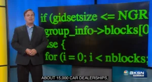 Screenshot of a news broadcast, with a newsanchor standing in front of a large screen showing closeup green text of computer code