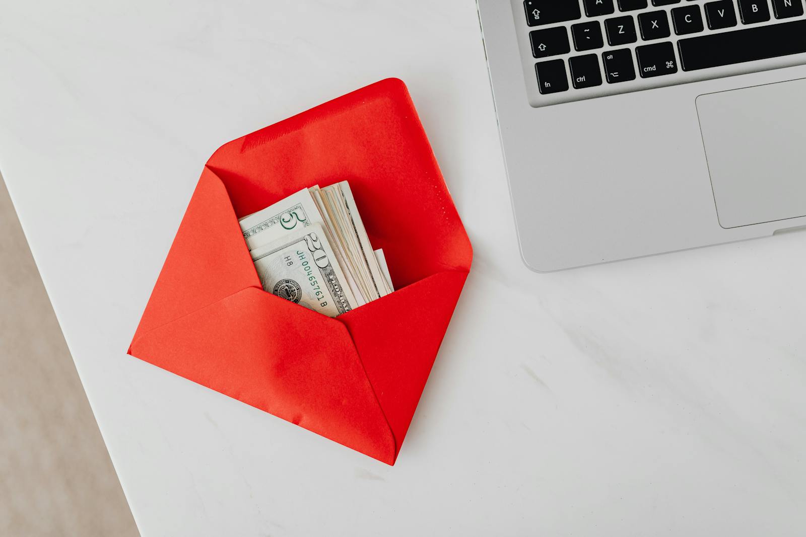 A red envelope filled with dollar bills next to a laptop on a marble desk.